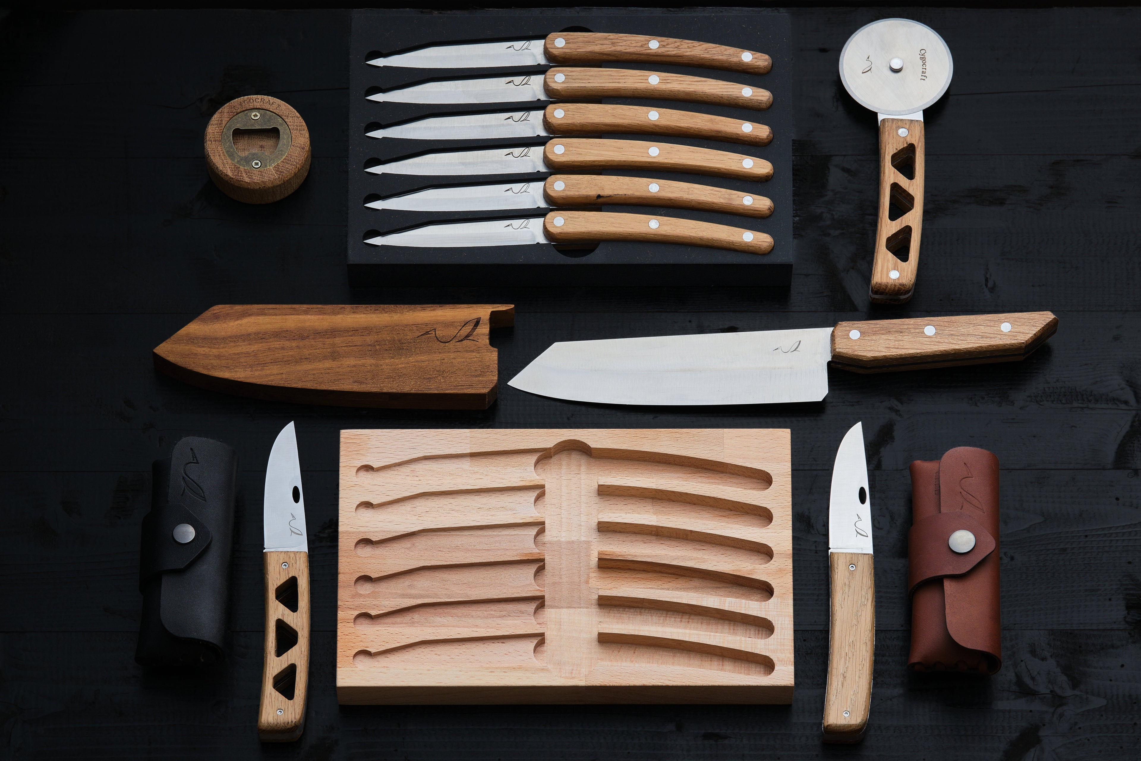 Ensemble de couteaux artisanaux disposés à plat sur un fond en bois noir : plusieurs couteaux à manche en bois, une grande lame de chef, des couteaux pliants avec étuis en cuir, un coupe-pizza, un ouvre-bouteille et des supports en bois, présentés de manière symétrique et élégante